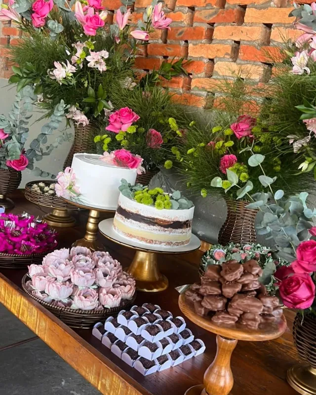 Mesa de festa com dois bolos, bandejas de docinhos e arranjos de flores rosa